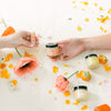 Two hands holding small jars of Fresh Pickins Farm Unscented organic body butter with flowers and scattered petals on a white surface